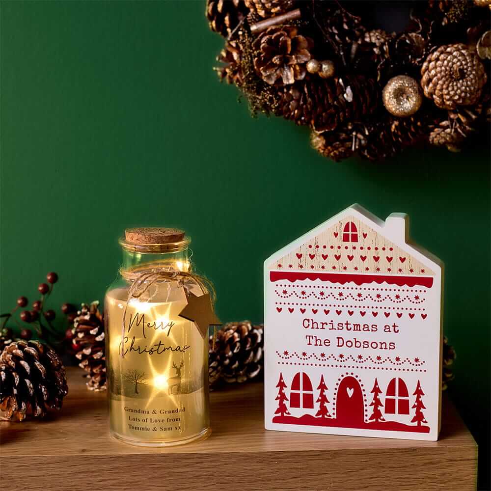 Personalised Christmas lantern and house-shaped decoration displayed on a wooden table with festive pinecones and a Christmas wreath. Personalised Christmas lantern and house-shaped decoration displayed on a wooden table with festive pinecones and a Christmas wreath.